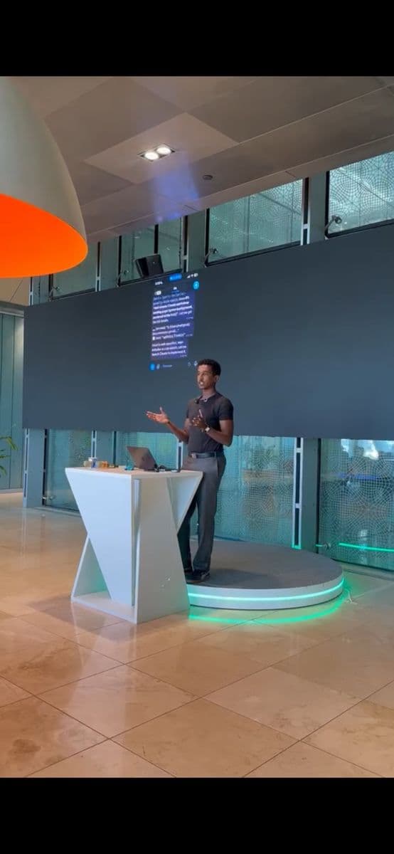 A presenter speaks on a lit stage in front of a large screen showing a chat on a phone.